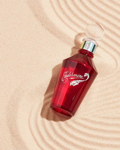 Red glass bottle with clear spherical stopper set on tan sand with concentric wave patterns and side lighting for visual appeal.