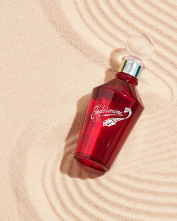 Red glass bottle with clear spherical stopper set on tan sand with concentric wave patterns and side lighting for visual appeal.