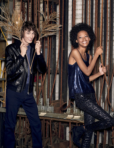 Two fashion models in a studio setting posing with gold decorative branches. A man in a leather jacket and a woman in a sequin top.