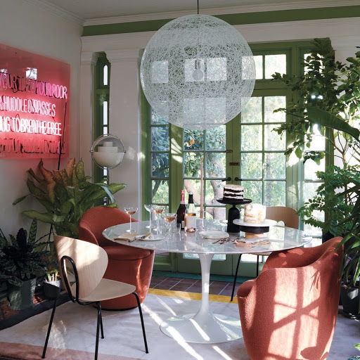 A dining room interior with a white marble table, surrounded by coral-colored chairs, illuminated by a globe pendant light.