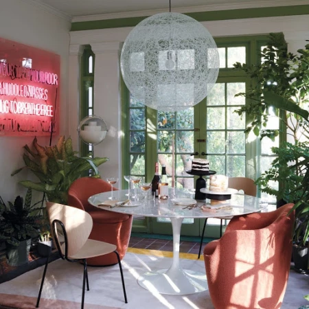A dining room interior with a white marble table, surrounded by coral-colored chairs, illuminated by a globe pendant light.