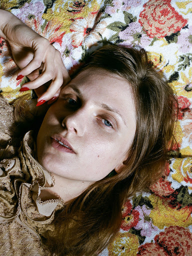 Woman with red nails lies on a floral patterned surface, gazing directly at the camera with neutral expression in a medium close-up studio portrait.