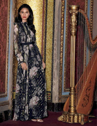Elegant woman in a floral maxi dress poses in front of a gilded wall with a harp, creating a vintage-inspired fashion photograph.