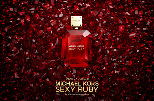 A red glass Michael Kors perfume bottle sits on a backdrop of faceted red gemstones, creating a visually striking beauty product shot.