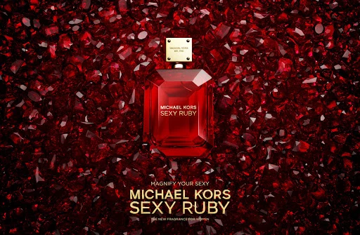 A red glass Michael Kors perfume bottle sits on a backdrop of faceted red gemstones, creating a visually striking beauty product shot.
