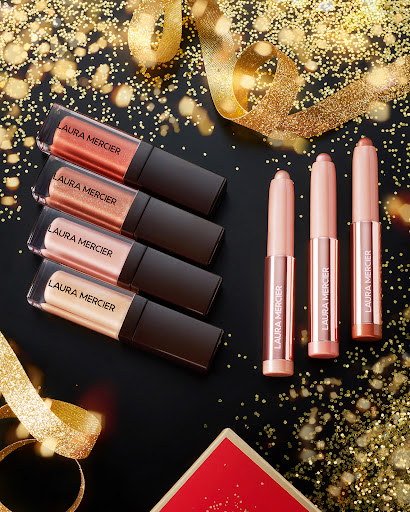 Laura Mercier makeup products including eyeshadow sticks and lip glosses arranged on a black surface with gold glitter and ribbons.