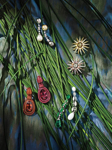 Elegant dangling earrings are displayed on a lush bed of green blades of grass set against a soft blue patterned backdrop.