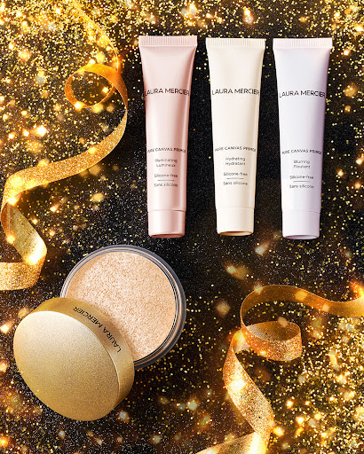 Laura Mercier makeup products, including primer tubes and a setting powder jar, are arranged on a glittering gold background.
