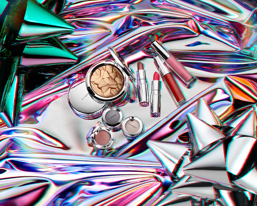 MAC cosmetics still life: shimmering powders, lipsticks, and lip glosses arranged against a holographic, iridescent background. Beauty aesthetic.