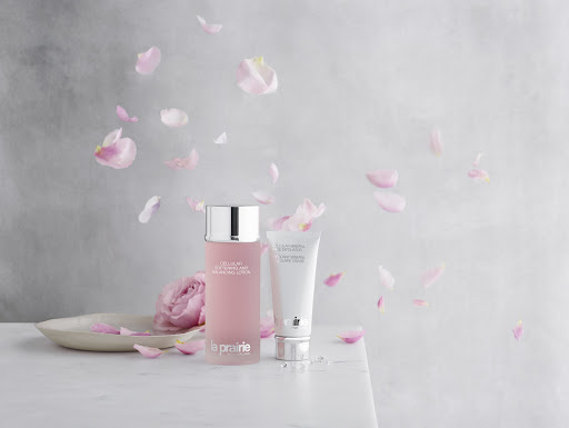 La Prairie skincare products, including a frosted pink bottle and white tube, are displayed with rose petals on a marble surface.