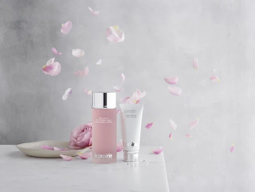 La Prairie skincare products, including a frosted pink bottle and white tube, are displayed with rose petals on a marble surface.