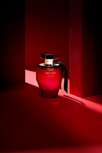 A red Victoria's Secret perfume bottle, Very Sexy Eau de Parfum, sits on a red surface with geometric red background shapes and shadows.