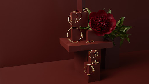 Elegant gold hoop earrings and studs displayed on red geometric platforms alongside a red flower. Jewelry and beauty product shot.