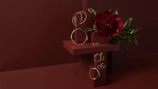 Elegant gold hoop earrings and studs displayed on red geometric platforms alongside a red flower. Jewelry and beauty product shot.