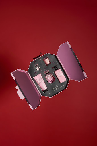 Flat lay photo of a pink gift set featuring a perfume bottle, body lotion, and candle, all arranged in an open box against a red backdrop.