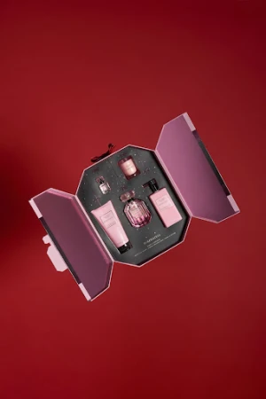 Flat lay photo of a pink gift set featuring a perfume bottle, body lotion, and candle, all arranged in an open box against a red backdrop.