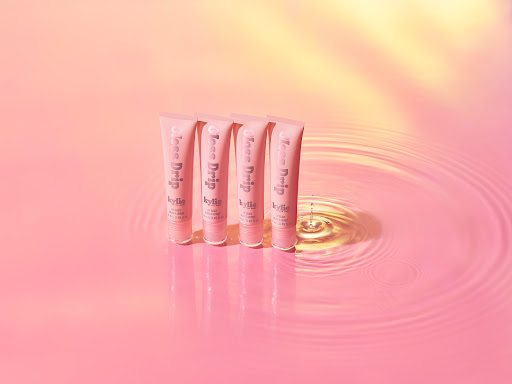 Kylie Cosmetics Gloss Drip lip gloss tubes arranged on a pink surface with water ripple effect, in a beauty and cosmetics product shot.