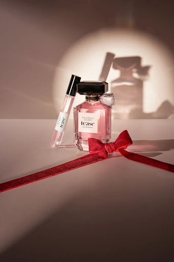 Still life of 'Tease' perfume bottle and rollerball with red bow, presented in soft, diffused studio lighting and elegant composition.
