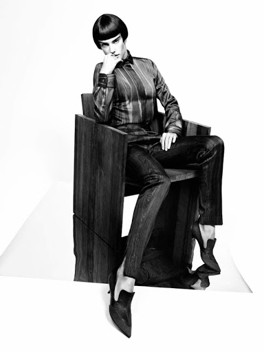 Elegant woman with short bowl haircut sits in a modern wooden chair wearing shiny dark striped shirt, dark pants, and stiletto heels in a photo studio.