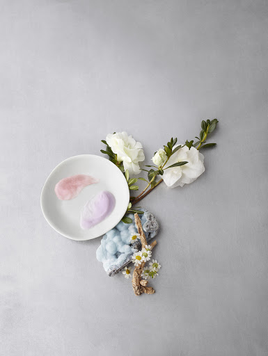 Elevated still life with white flowers, white plate holding cosmetic creams, mineral stone and white daisies on a grey background.