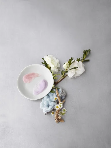 Elevated still life with white flowers, white plate holding cosmetic creams, mineral stone and white daisies on a grey background.