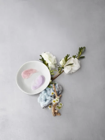 Elevated still life with white flowers, white plate holding cosmetic creams, mineral stone and white daisies on a grey background.