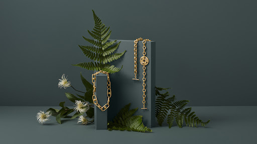 Still-life shot of gold bracelets displayed on a grey block, accented with green fern leaves and small white flowers, against a blue backdrop.