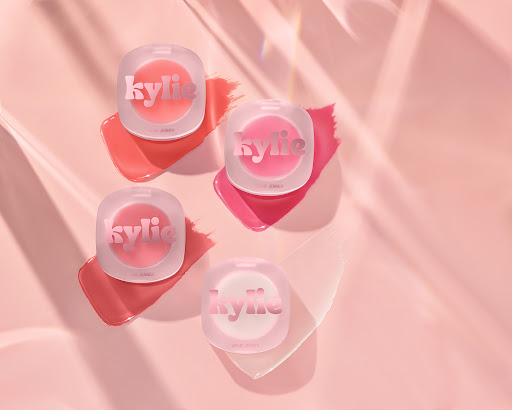 Four Kylie Cosmetics lip glosses sit on a pink surface, casting shadows and highlighted by diffused lighting for a beauty product shot.