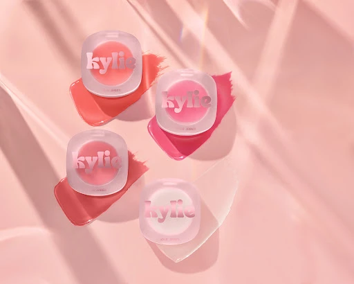 Four Kylie Cosmetics lip glosses sit on a pink surface, casting shadows and highlighted by diffused lighting for a beauty product shot.