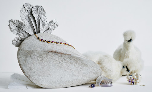Jewellery still life featuring a gold necklace with gemstones, rings, and earrings next to two white Silkie chickens on a white surface.