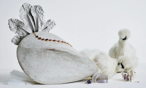 Jewellery still life featuring a gold necklace with gemstones, rings, and earrings next to two white Silkie chickens on a white surface.