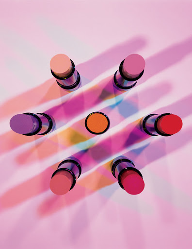 Overhead shot of colorful lipsticks arranged in a starburst pattern on a pink background with diffused light and shadows creating visual interest.