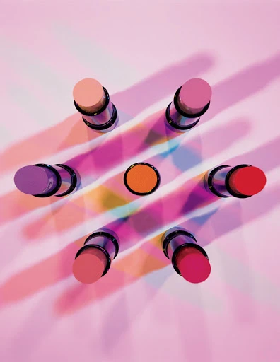 Overhead shot of colorful lipsticks arranged in a starburst pattern on a pink background with diffused light and shadows creating visual interest.