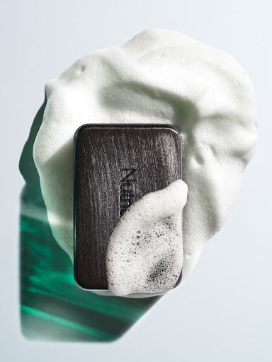 Studio shot of dark-colored Neutrogena soap bar on white surface, surrounded by white soap suds, and reflecting green and white light.