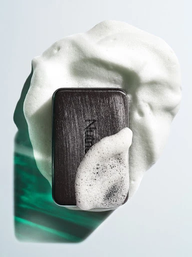 Studio shot of dark-colored Neutrogena soap bar on white surface, surrounded by white soap suds, and reflecting green and white light.