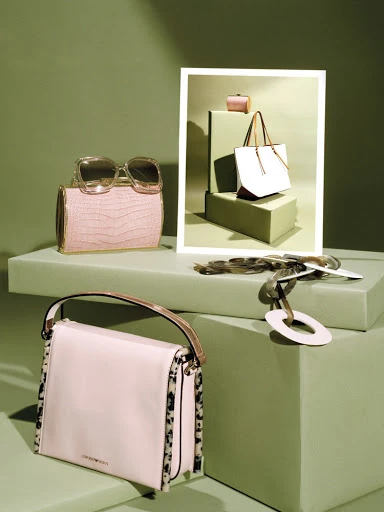 Still life shot featuring luxury handbags and sunglasses arranged on light-green platforms against a matching background, product photography.