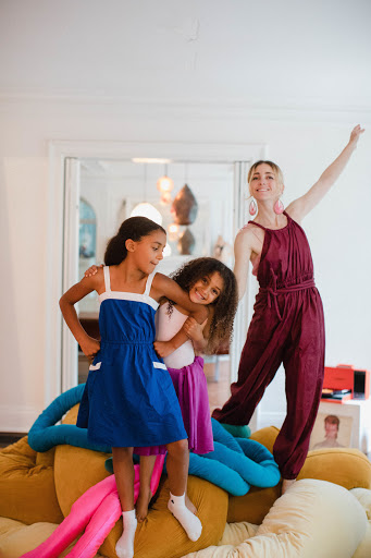 Two children embrace on a beanbag, while an adult woman stands behind them with arms raised in a bright, naturally lit home interior.