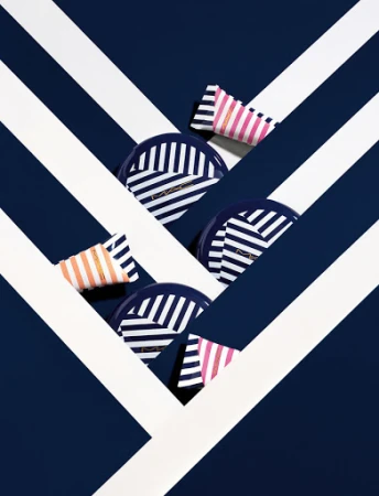Cosmetic compacts and tubes with navy and white stripes arranged on dark blue background with white diagonal stripes