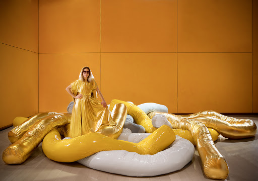 Woman in gold dress poses on inflatable yellow and white tubes in a room with a yellow wall, creating a striking fashion statement.