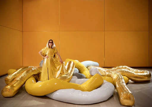 Woman in gold dress poses on inflatable yellow and white tubes in a room with a yellow wall, creating a striking fashion statement.