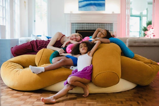 Three females relax on a large beanbag in an indoor setting, with a fireplace and pink floral accents in the background.