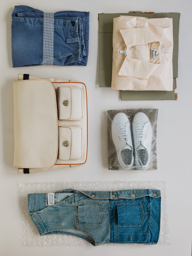 Flat lay of a neatly packed cream-colored purse, folded blue jeans, white sneakers in bubble wrap, and a folded light pink shirt.