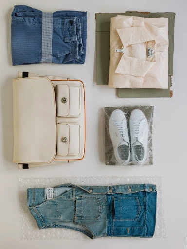 Flat lay of a neatly packed cream-colored purse, folded blue jeans, white sneakers in bubble wrap, and a folded light pink shirt.