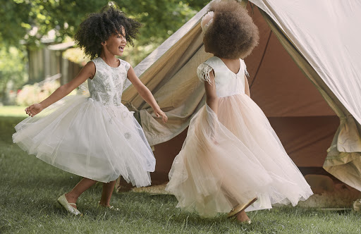 Two young girls in formal dresses are joyfully playing near a tent in a grassy outdoor setting, exuding a playful and lighthearted mood.
