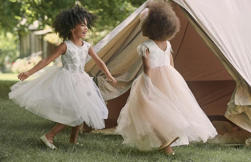 Two young girls in formal dresses are joyfully playing near a tent in a grassy outdoor setting, exuding a playful and lighthearted mood.