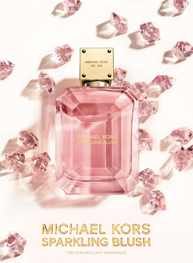 Michael Kors Sparkling Blush perfume bottle against a white backdrop with scattered gem-like objects, promoting fragrance.