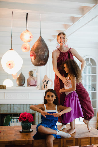 An adult woman standing with two girls in a home setting, featuring interior design elements like pendant lights and wooden furniture.