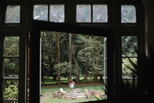 Young child stands on outdoor table surrounded by tree stump chairs in a forest clearing, viewed through a window frame.
