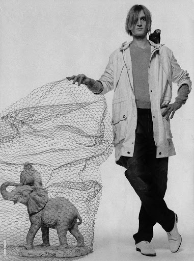 Monochrome portrait of a man with a bird on his shoulder standing next to an elephant statue covered in netting against a white backdrop.