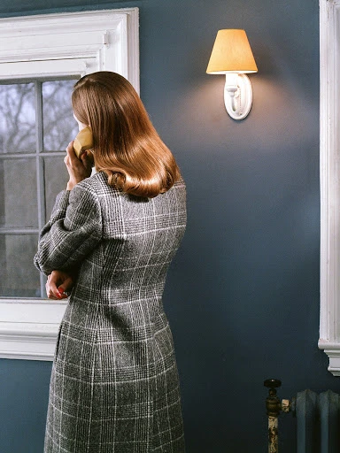 Woman in a patterned grey coat stands indoors near a window while holding a landline telephone receiver to her ear, bathed in warm, lamp lighting.